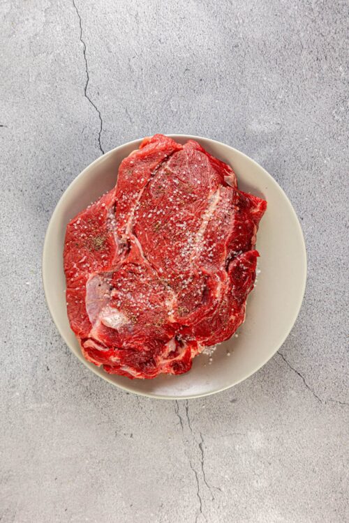 Fresh raw beef chuck roast seasoned with salt and pepper on a white plate.