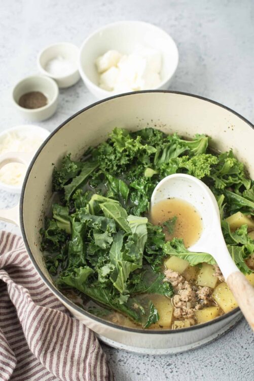 Fresh kale and potato soup cooking in a pot with seasoning ingredients in the background.