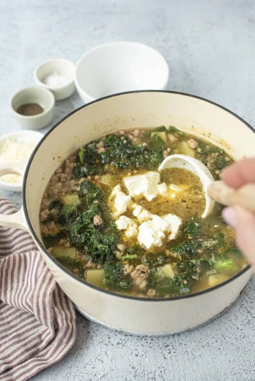 Comforting homemade soup with greens, ground meat, and crumbled cheese in a white pot.