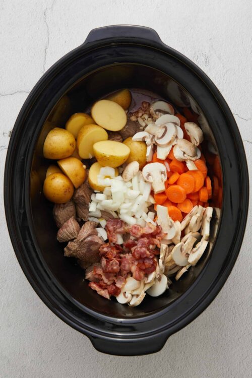 Fresh ingredients in a slow cooker for beef stew with potatoes, carrots, mushrooms, onions, and bacon.