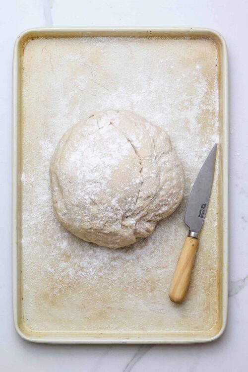 Flour-dusted bread dough on a baking sheet with a bench scraper.