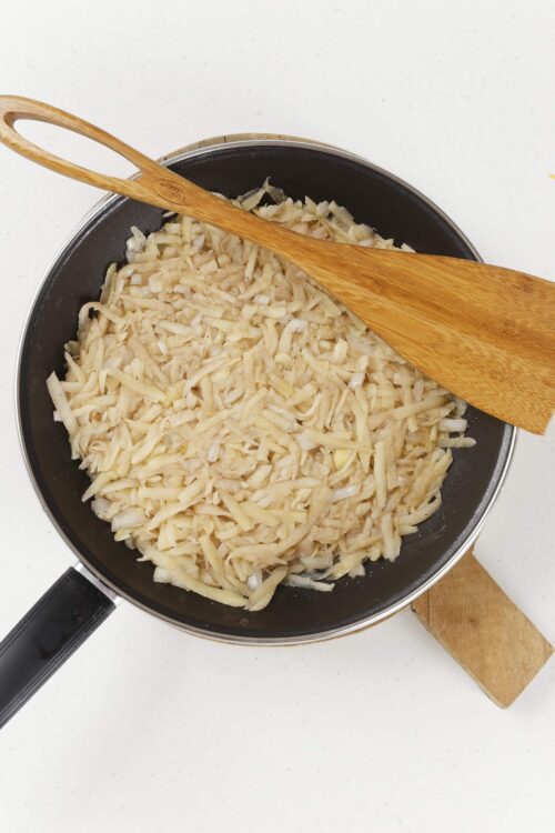 Sautéed chopped onions cooking in a black skillet with a wooden spatula on white countertop.