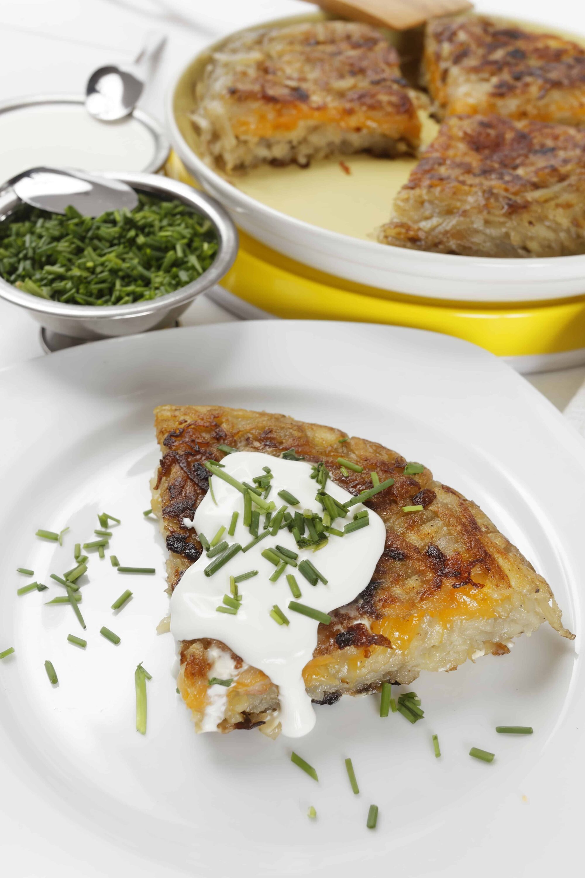 Creamy cheesy potato casserole with sour cream and green chives on top, served with a side of chopped herbs.