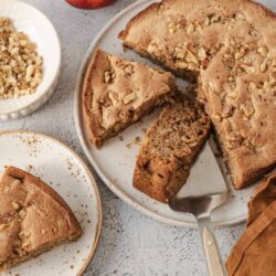 Moist apple nut cake with chopped walnuts on a white plate, fresh apples in background.