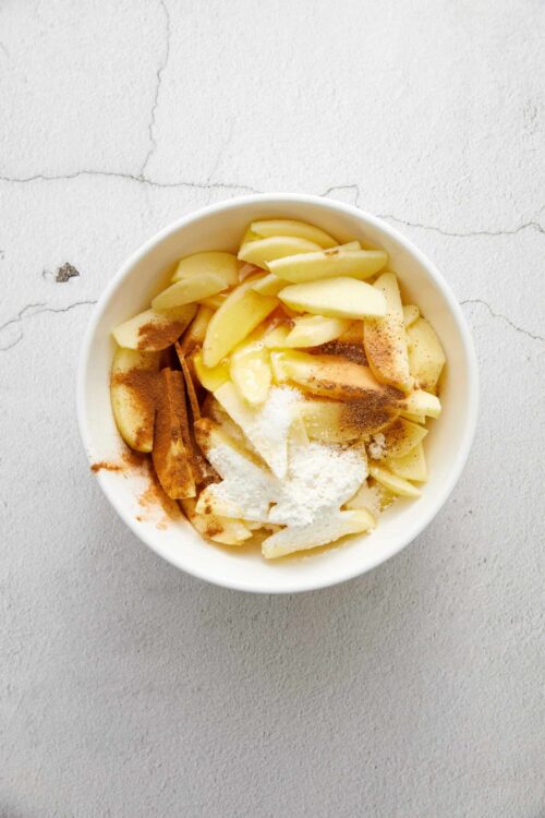 Sliced apples with cinnamon, sugar, and flour in a white bowl for baking or cooking.