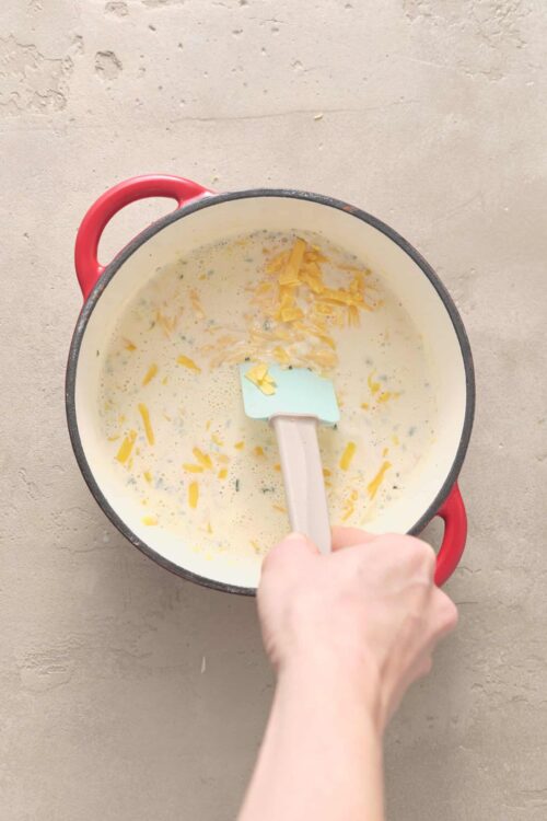 Creamy cheese soup with shredded cheddar, being stirred in a red cast iron pot.
