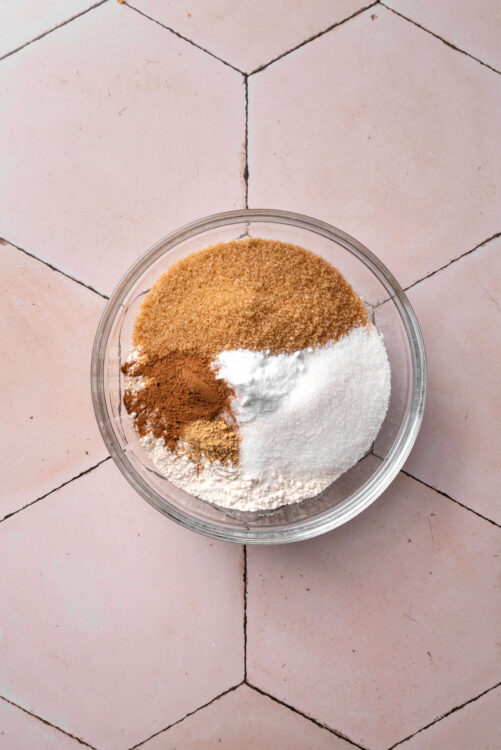 Ground cinnamon, sugar, and spices in a glass bowl on pink tiled kitchen floor.