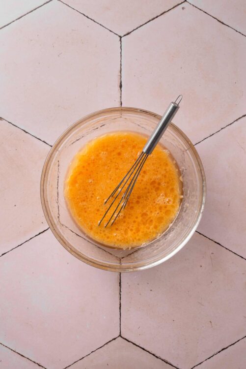 Bright orange beaten eggs in a glass bowl with a whisk, on a pink tiled kitchen counter.