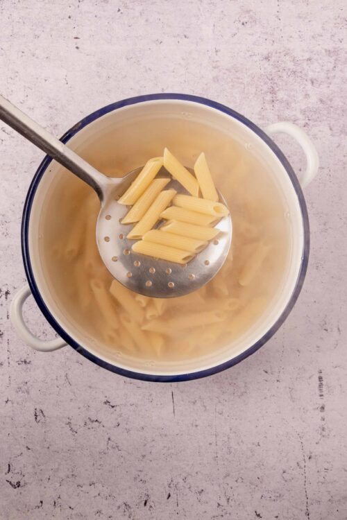 Pasta being cooked in a white pot with a metal slotted spoon holding penne pasta.