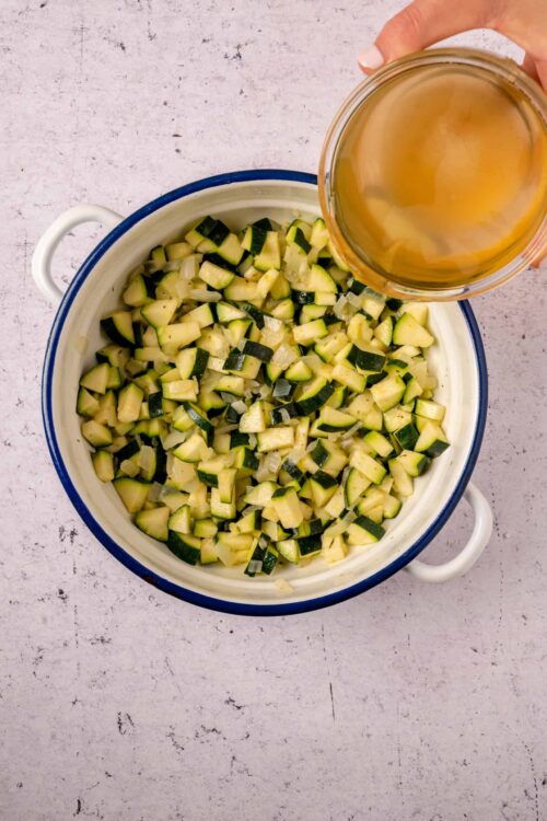 Diced zucchini and onions in a white ceramic dish with a blue rim, hot broth being poured over vegetables.