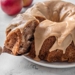 Golden apple cinnamon cake with caramel frosting on a white plate with fresh apples in the background.