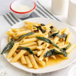 Pasta with sage and butter sauce on a white plate, side of Parmesan cheese, rustic table setting.