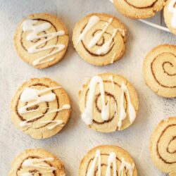 Buttery cinnamon roll cookies with white glaze drizzle on a white surface, perfect for dessert or snack.