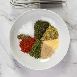 Finely ground herbs and spices on a white plate with a whisk on a marble countertop.