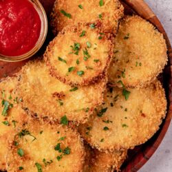 Golden breaded chicken cutlets with chopped parsley and a side of marinara sauce in a wooden bowl.
