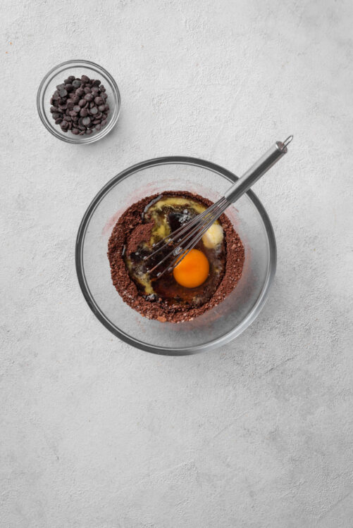 Chocolate baking ingredients in a glass mixing bowl with a whisk, egg, butter, and cocoa powder on a white textured surface.