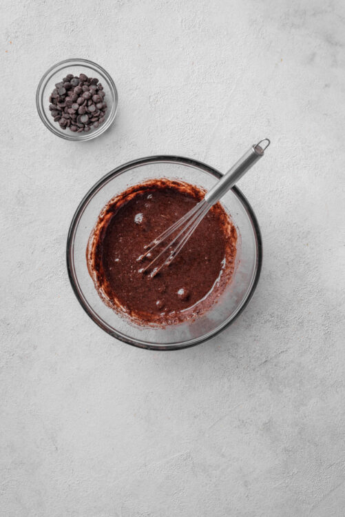 Chocolate cookie dough mixture in a glass bowl with a whisk, with chocolate chips in a small bowl nearby.