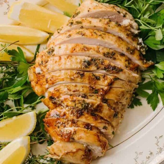 Juicy grilled chicken breast with herbs and lemon wedges on a white platter.