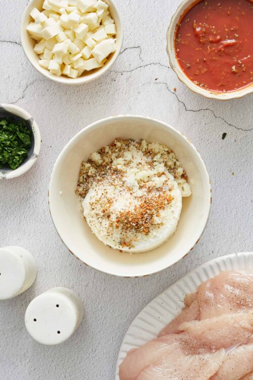 Cream cheese and shredded cheese for chicken casserole, with spices and diced cheese, on a white textured surface.