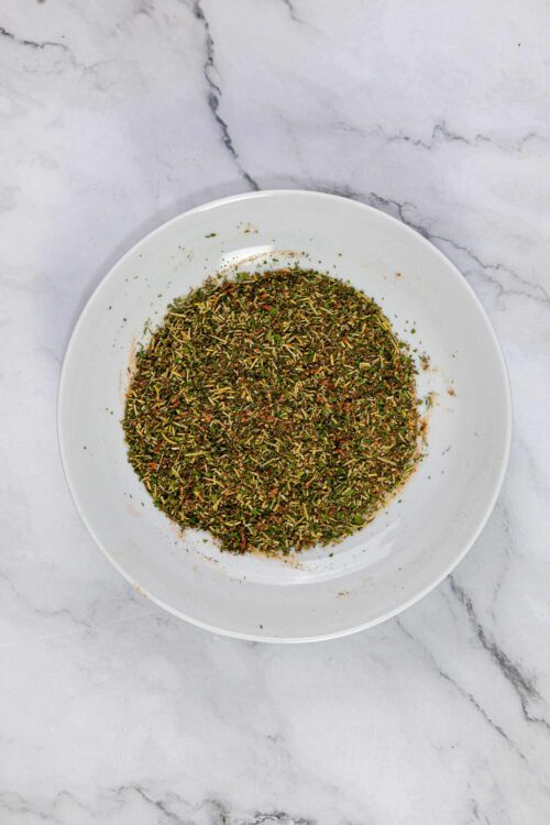 Dried Italian herb seasoning mixture in a white bowl on marble surface.