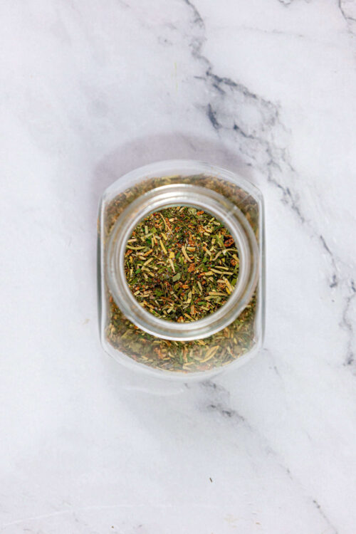 Dried herb spice mix in glass jar on white marble surface for baking and cooking.