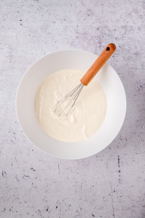 Creamy white batter in a white mixing bowl with a whisk and wooden handle for baking recipes.