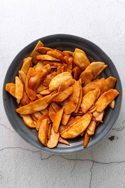 Sliced baked apples with cinnamon in a gray bowl on textured surface, perfect for apple desserts or snacks.
