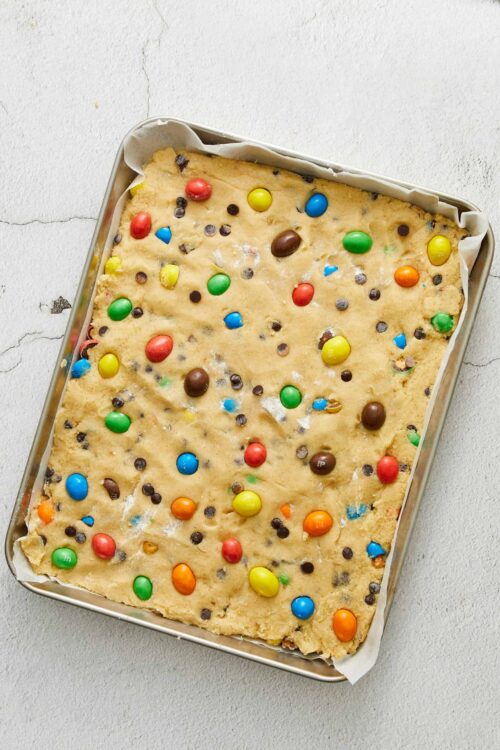 Colorful candy-coated cookie bar in a baking pan, topped with M&Ms and chocolate chips.