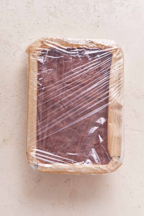 Freshly baked chocolate brownie covered with plastic wrap on a baking tray.