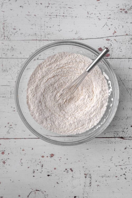 Flour in a glass mixing bowl with a whisk on a rustic white wooden background.