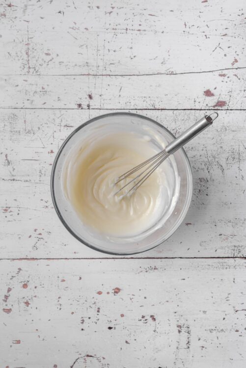 Creamy homemade whipped cream in a glass bowl with a whisk, on a rustic white wooden surface.