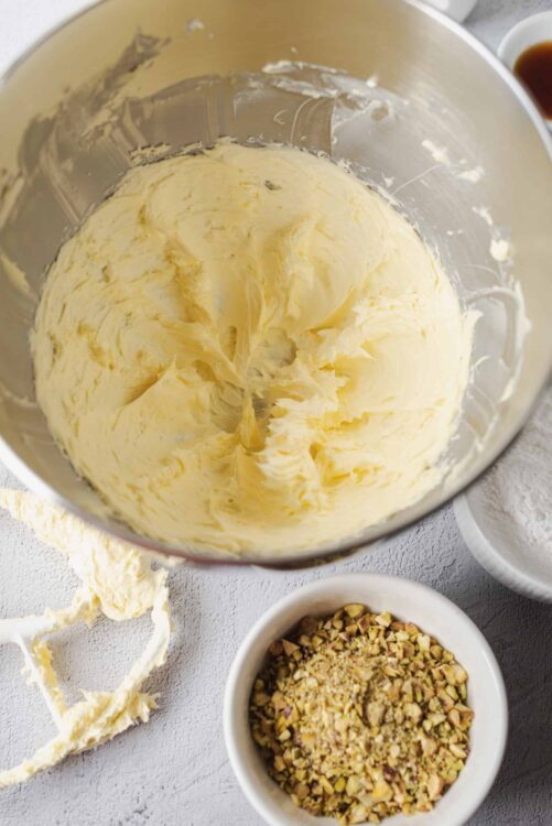Creamy butter mixture in mixing bowl with chopped pistachios on white surface.