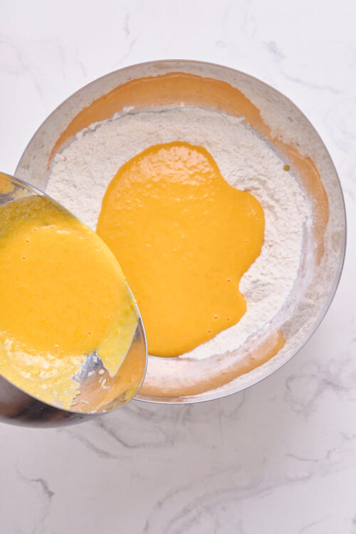 Light yellow cake batter being poured into flour mixture in a mixing bowl.