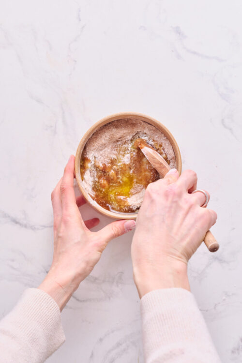 Creamy caramel sauce being stirred in a small bowl with a wooden spoon on a white marble surface.