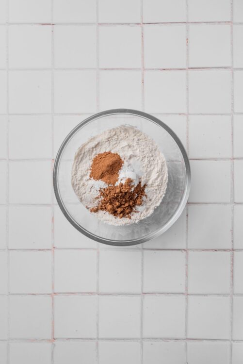 Flour and cocoa powder in a glass mixing bowl on a white tiled surface.