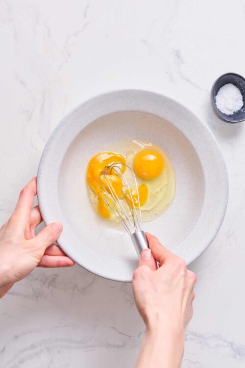 Whisking eggs in a white bowl for baking, with salt on a small dish, on a clean white marble surface.