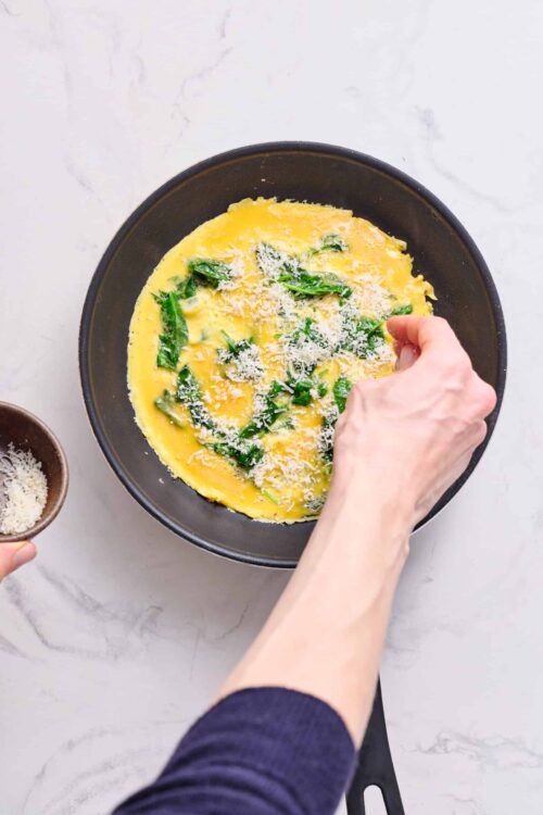 Freshly beaten eggs with spinach and grated cheese in a black skillet cooking for breakfast or recipe preparation.