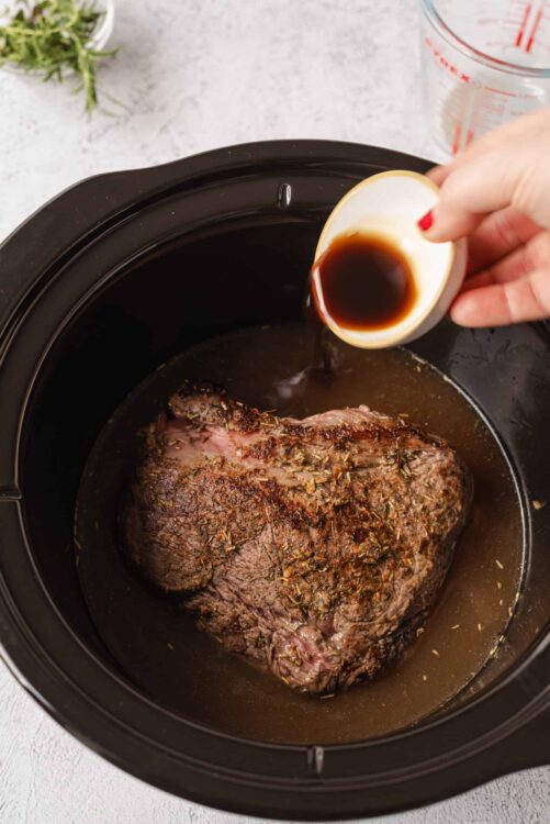Juicy beef pot roast being slow-cooked in a crockpot with broth and herbs.