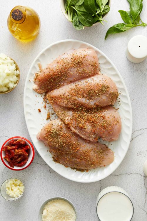 Raw chicken breasts seasoned with herbs and spices on a white platter.