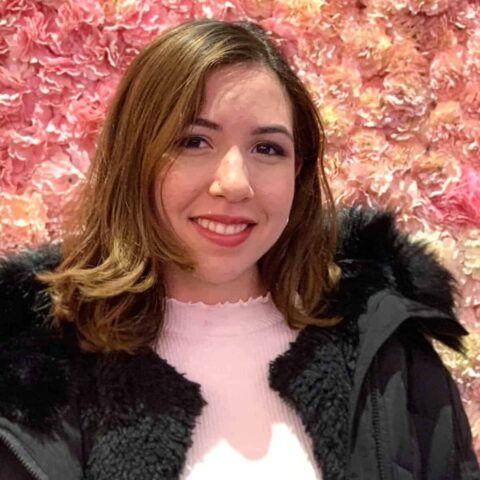 Fresh-faced woman smiling in front of pink flower wall, casual fall outfit, cozy black jacket and pink top, bright and cheerful ambiance.