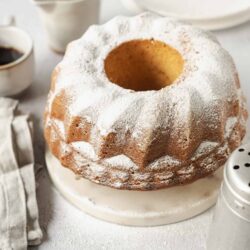 Fluffy vanilla bundt cake dusted with powdered sugar on a white cake stand. Perfect for dessert or special occasions.