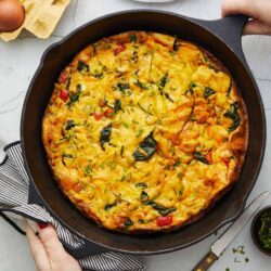 Savory vegetable and cheese frittata in a cast iron skillet on a white countertop with eggs and plates nearby.