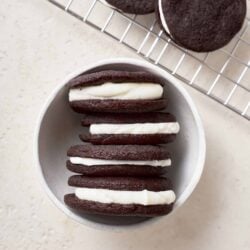 Soft chocolate sandwich cookies with vanilla cream filling on a white ceramic bowl and cooling rack.