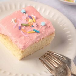 Sweet vanilla cake square with pink frosting and colorful sprinkles, served on a white plate.