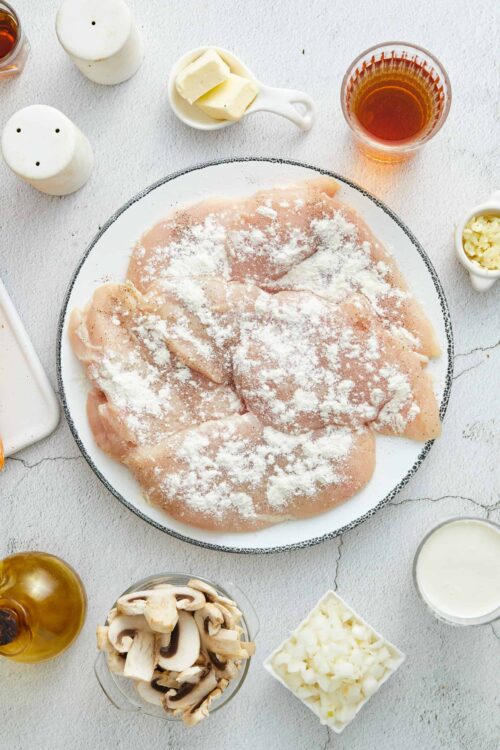 Fresh raw chicken breast sprinkled with flour, surrounded by mushrooms, butter, milk, and seasonings for homemade chicken cooking.