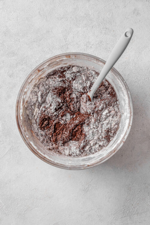 Rich chocolate frosting being mixed in a glass bowl with a white spatula.