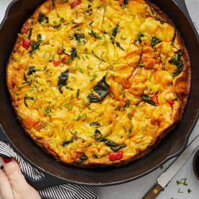 Savory vegetable and cheese frittata in a cast iron skillet on a white countertop with eggs and plates nearby.
