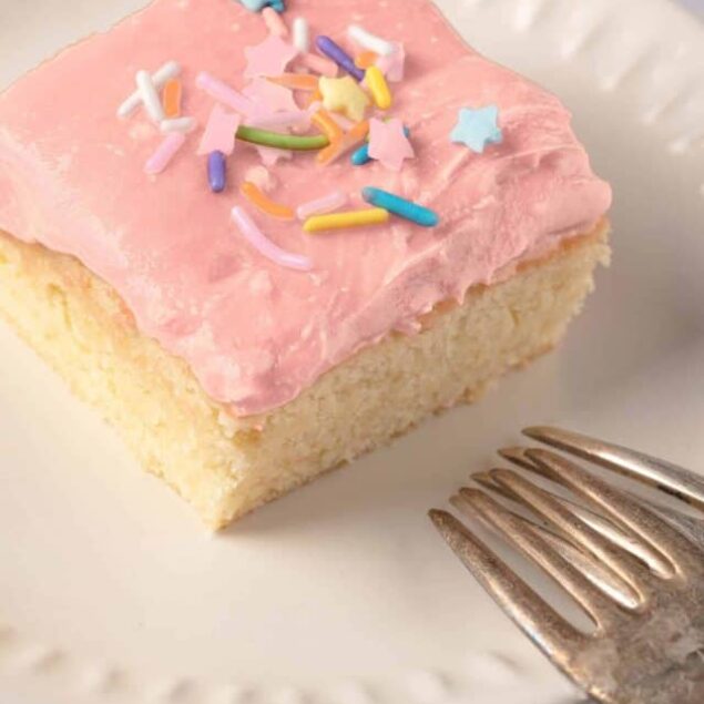 Sweet vanilla cake square with pink frosting and colorful sprinkles, served on a white plate.