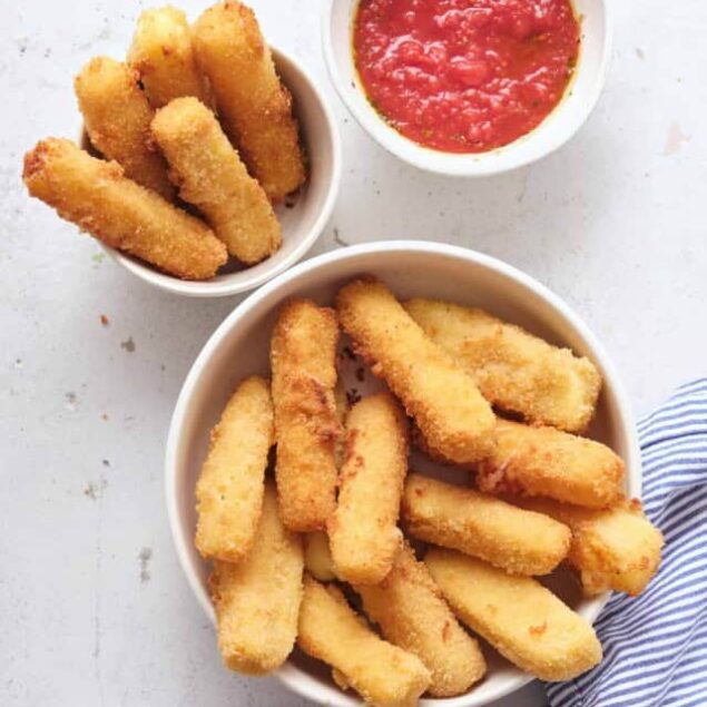 Crispy mozzarella sticks with marinara dipping sauce on a white plate and side bowl, served with a blue striped napkin.