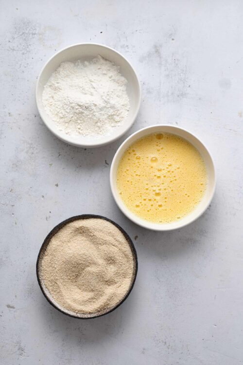 Flour, eggs, and breadcrumbs in bowls for baking on a white surface.